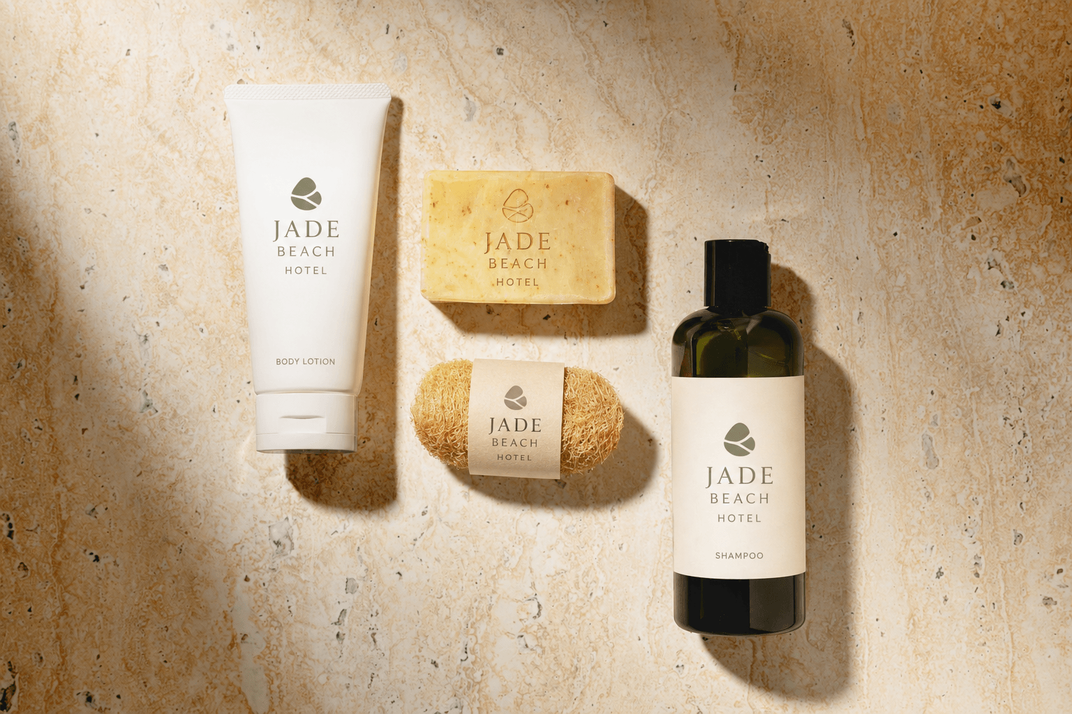Flat-lay of Jade Beach Hotel branded bathroom amenities: body lotion tube, soap bar, loofah, and shampoo bottle on travertine stone